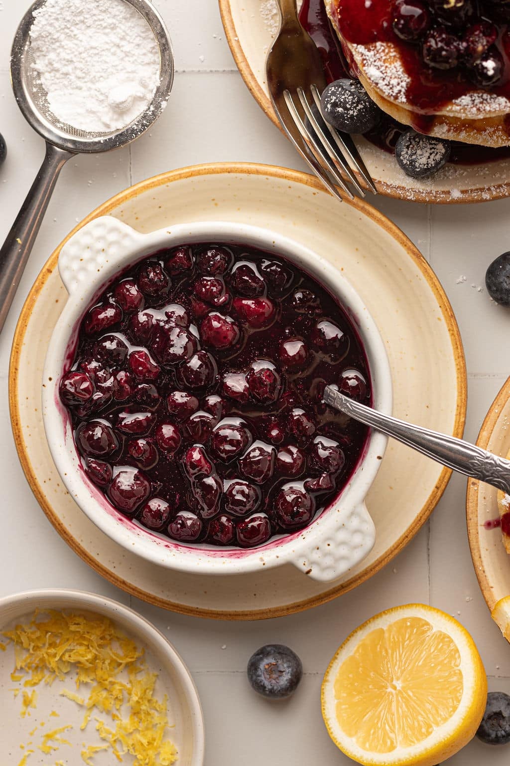 A close-up of a bowl filled with glossy blueberry compote, with a spoon resting inside. Surrounding the bowl are lemon zest, fresh blueberries, and a sifter with powdered sugar.