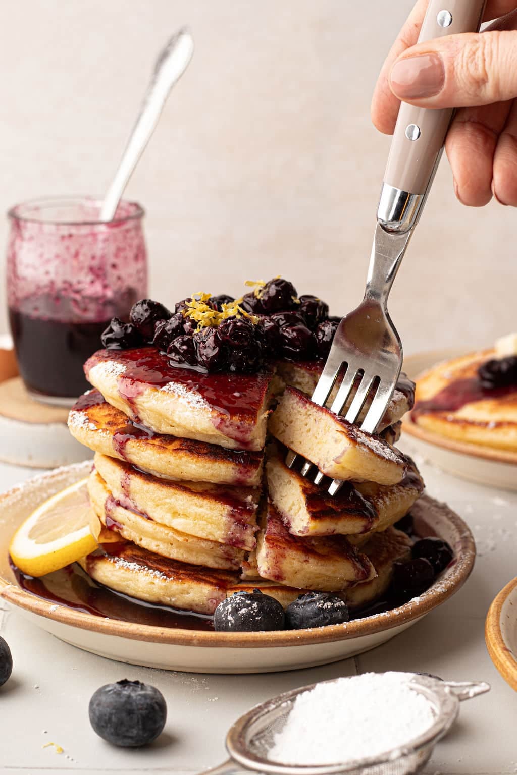 A hand cutting into a stack of pancakes, revealing the soft, fluffy interior. The pancakes are covered in blueberry compote, lemon zest, and powdered sugar. A jar of blueberry sauce and a second plate of pancakes are in the background.