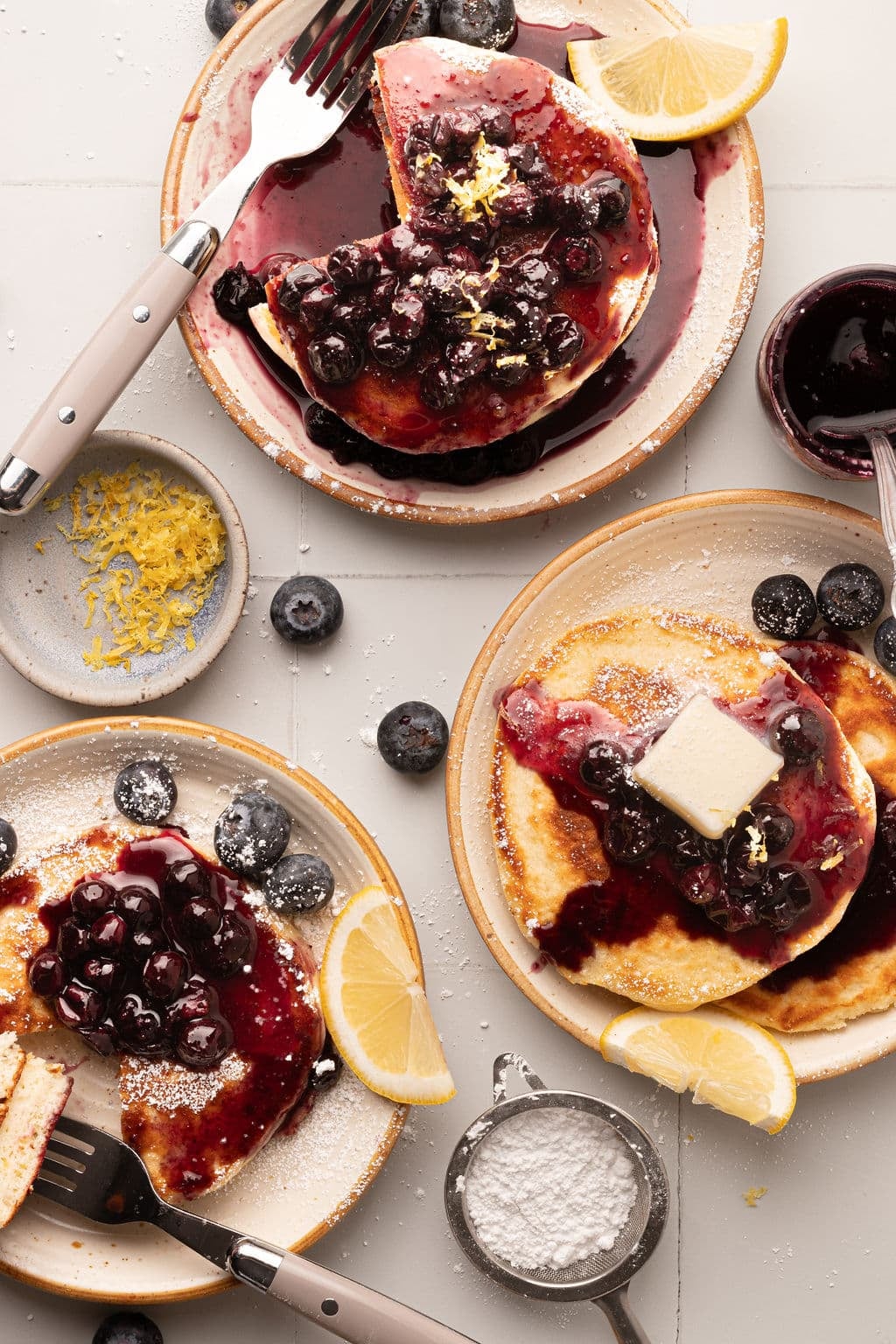 Three plates of pancakes with different levels of toppings—one with a butter pat melting over the blueberry compote, another with powdered sugar, and one partially eaten. Lemon slices, a sieve of powdered sugar, and fresh blueberries are scattered across the table.