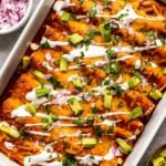 white casserole dish filled with red enchiladas topped with a drizzle of sour cream, chopped avocado, and cilantro