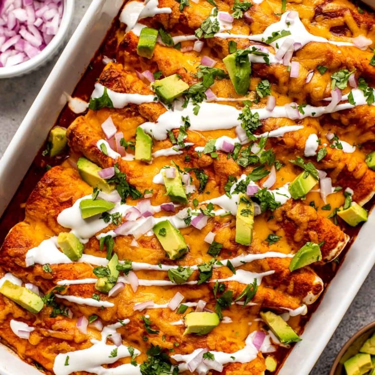 white casserole dish filled with red enchiladas topped with a drizzle of sour cream, chopped avocado, and cilantro