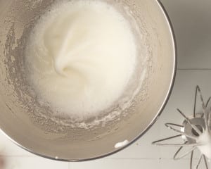 A close-up of a metal mixing bowl containing stiffly whipped egg whites, with a whisk attachment resting nearby.