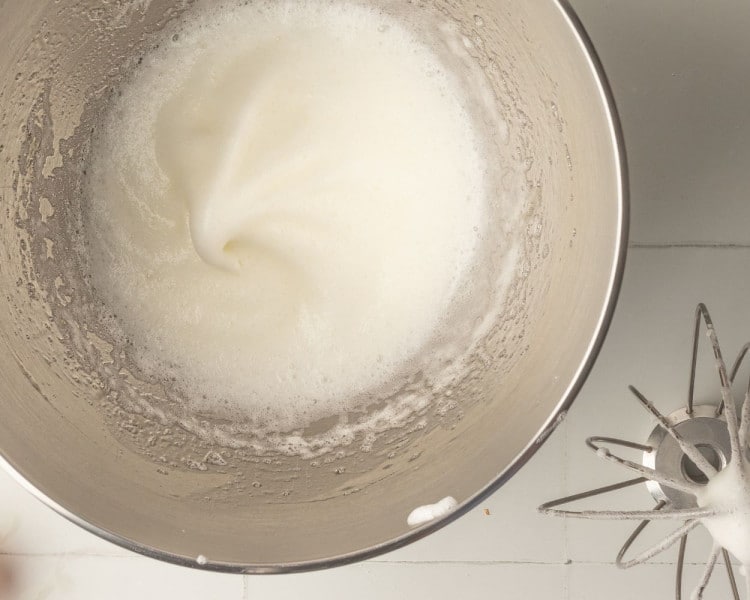 A close-up of a metal mixing bowl containing stiffly whipped egg whites, with a whisk attachment resting nearby.