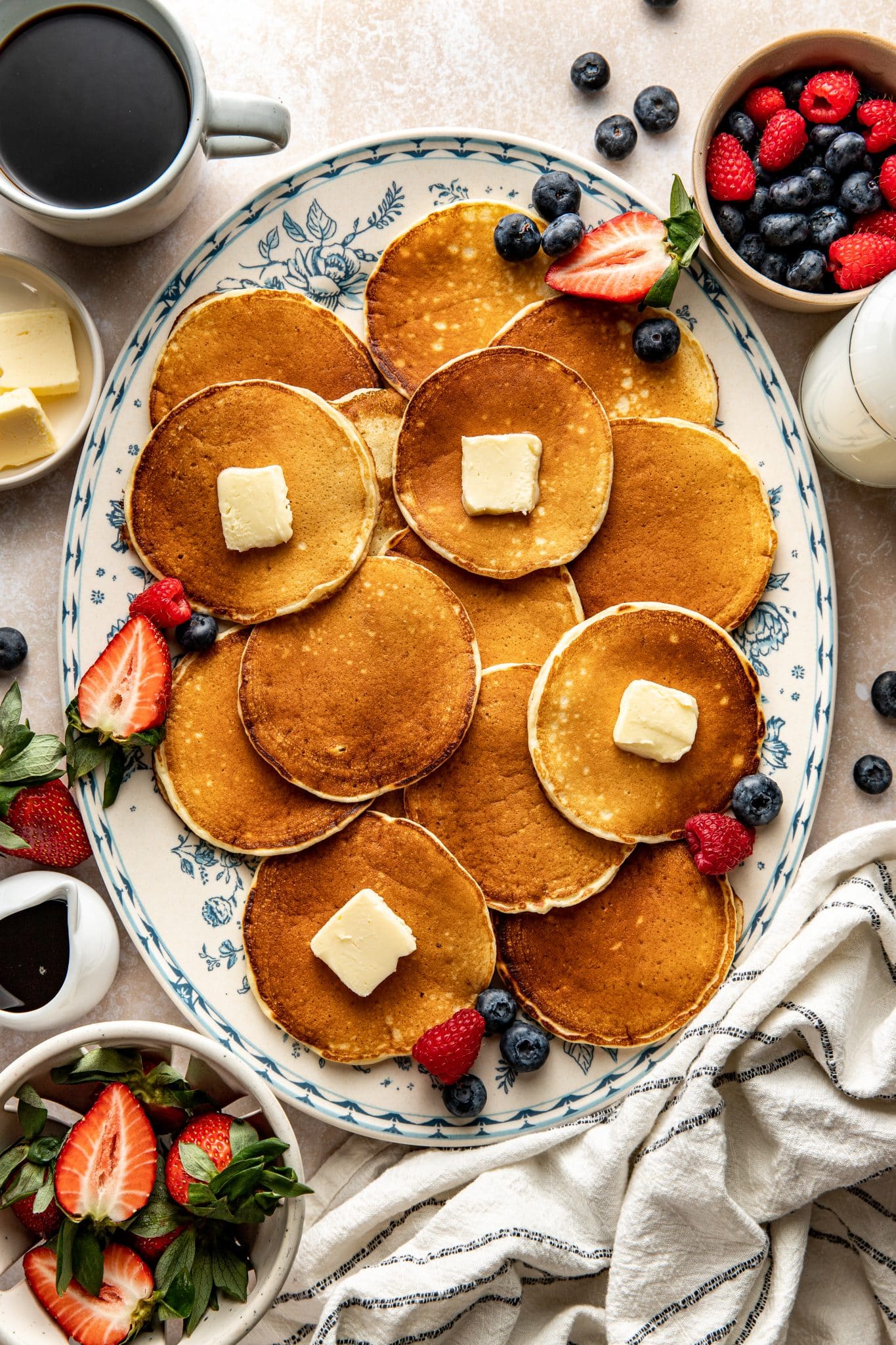 Overhead view of a large oval platter filled with golden buttermilk pancakes topped with pats of butter, surrounded by fresh strawberries, blueberries, a cup of coffee, a bowl of butter, and a bowl of mixed berries.