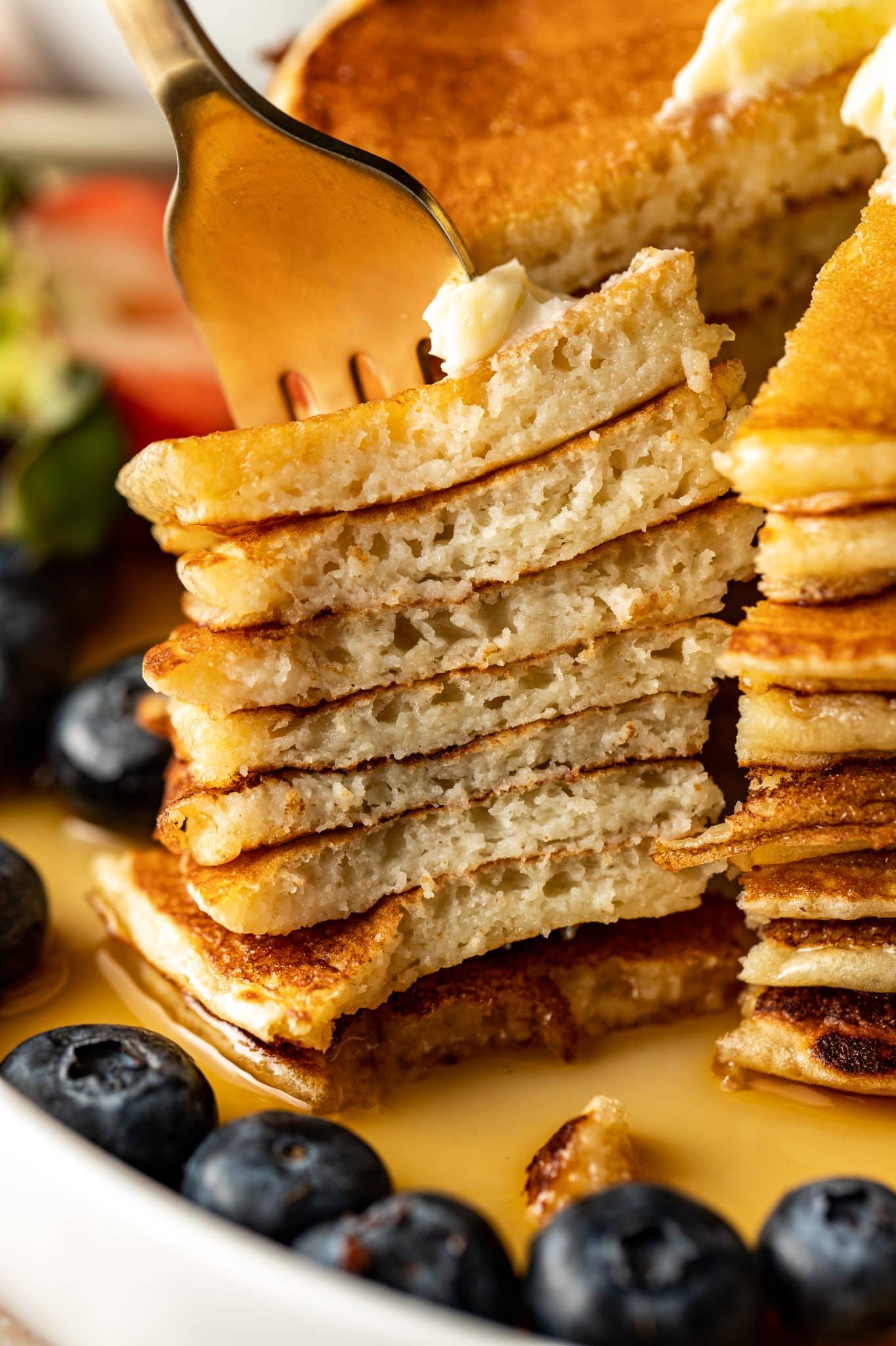Fork lifting a bite of stacked pancakes with visible fluffy texture and syrup pooling on the plate, with fresh berries scattered around.