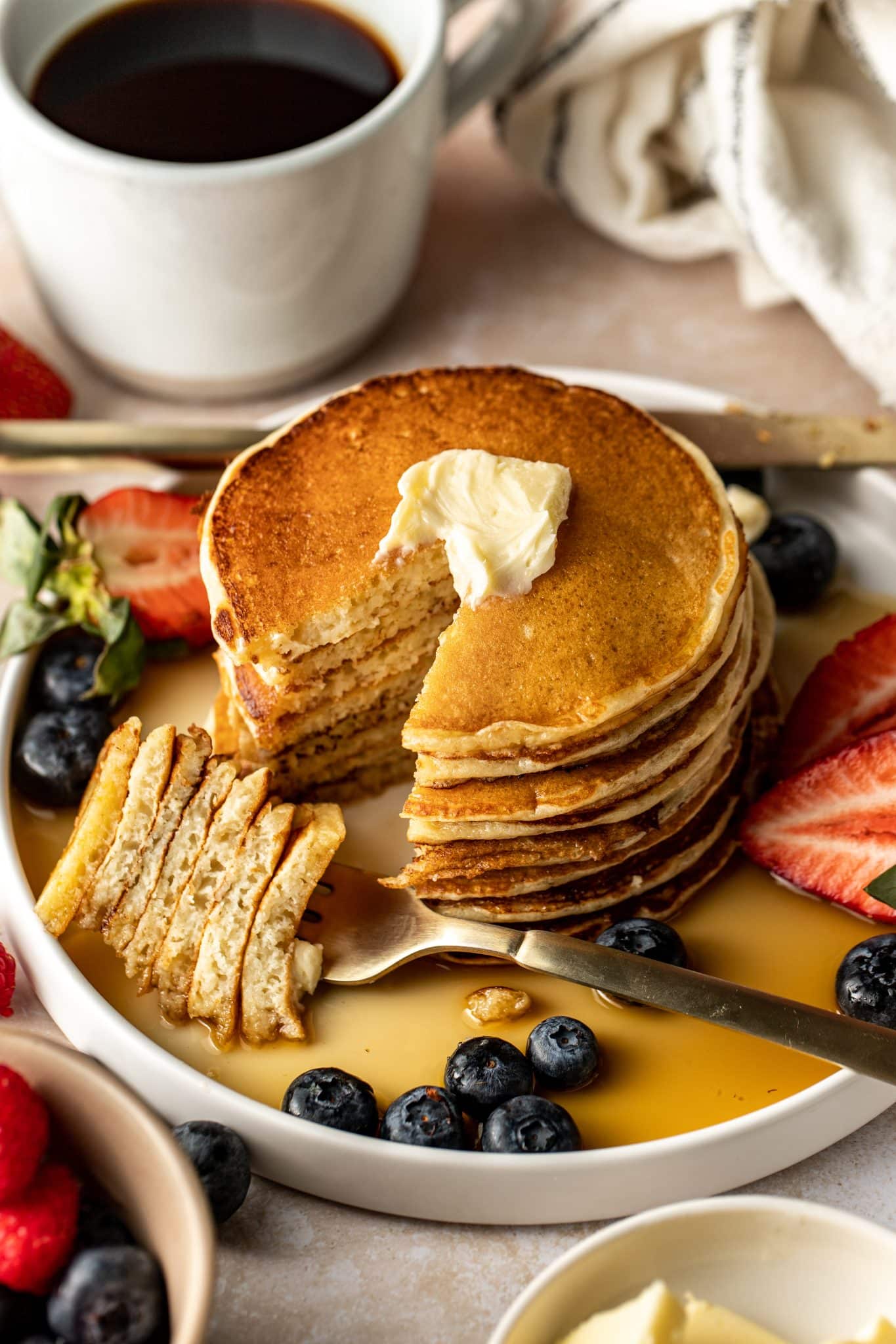 Partially eaten stack of pancakes topped with butter, with a fork holding a tall bite of sliced pancakes, surrounded by blueberries, strawberries, and syrup.