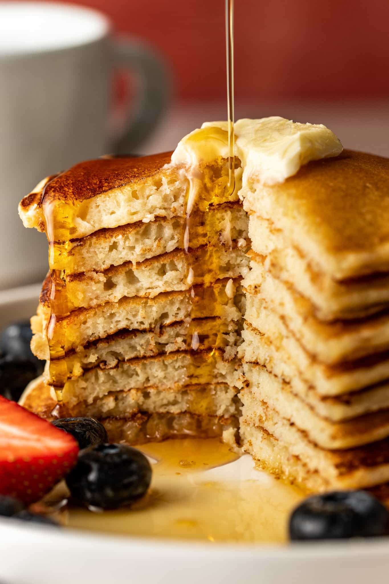 Maple syrup dripping over a tall, cut-open stack of pancakes topped with melting butter, revealing a tender and fluffy interior.