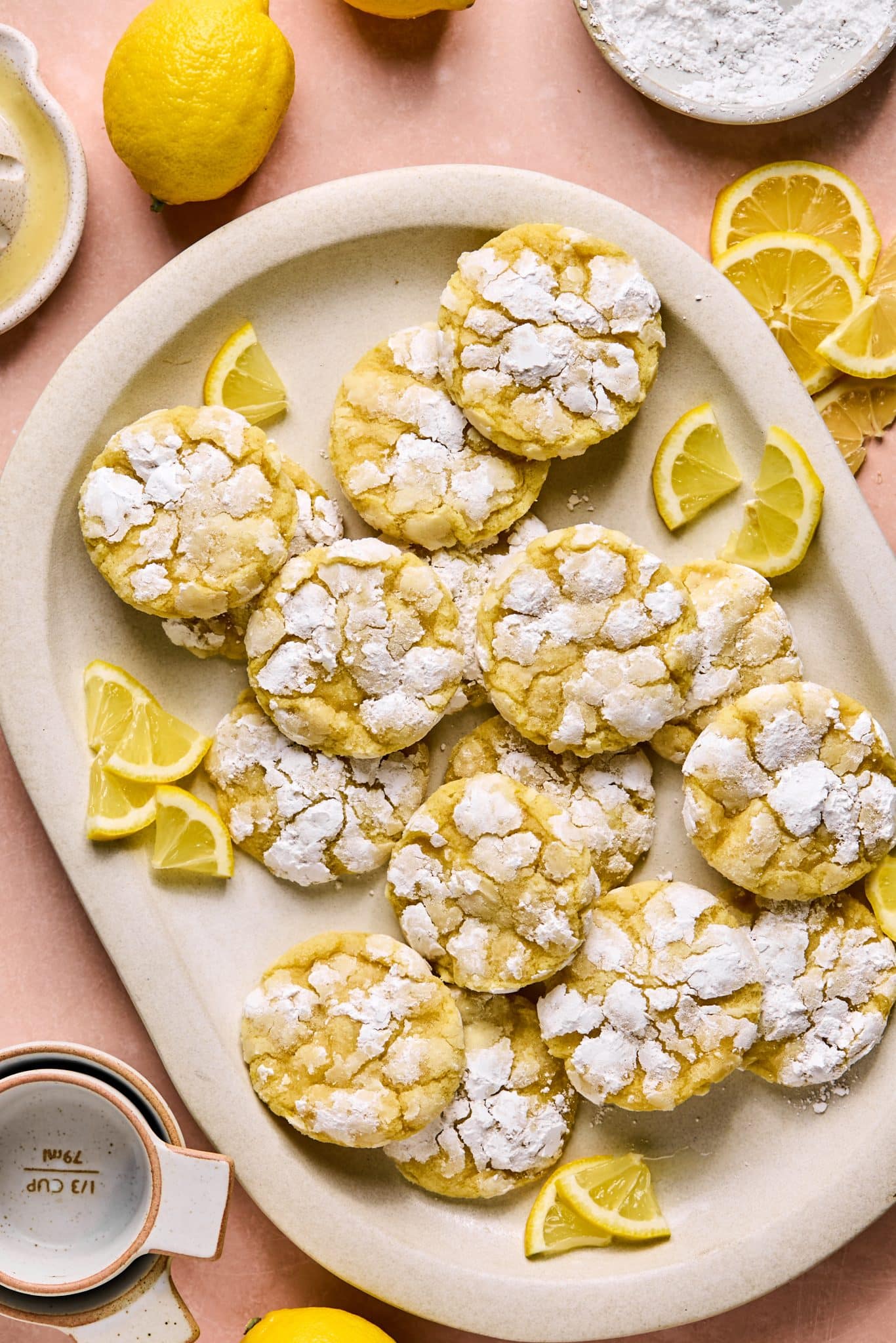 A large batch of lemon crinkle cookies on an oval beige platter, dusted with powdered sugar and surrounded by fresh lemon wedges on a peach surface.

