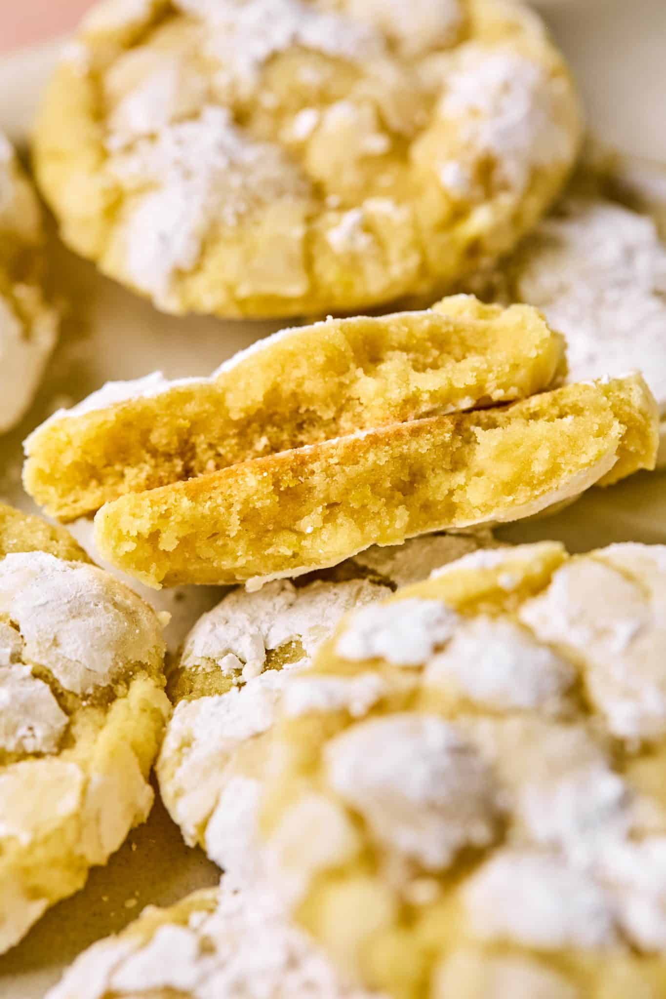 A lemon crinkle cookie broken in half to show the tender, moist center and golden edges, with crinkled powdered sugar tops and more cookies in the background.

