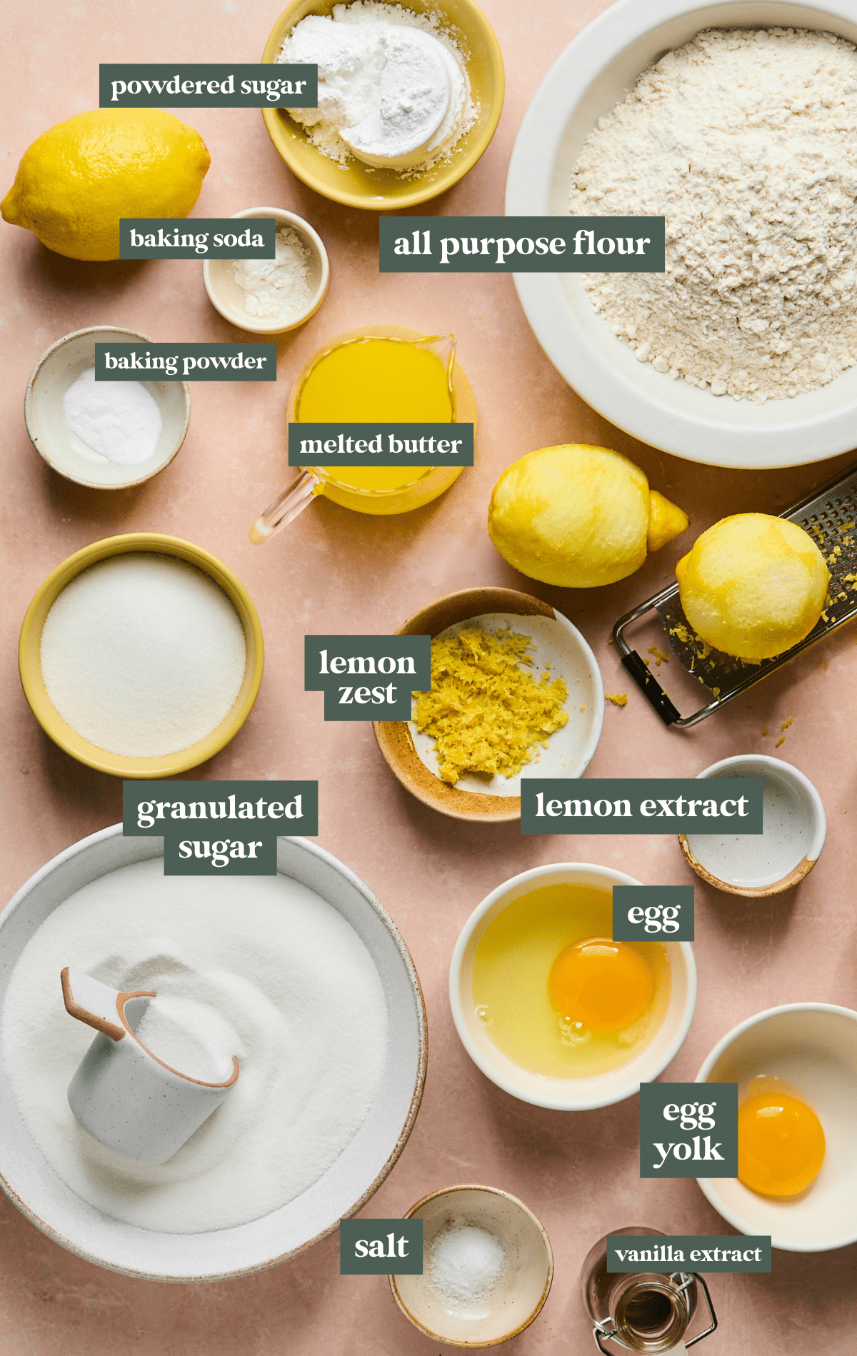 Flatlay of lemon crinkle cookie ingredients on a peach-colored surface. Includes all-purpose flour, powdered sugar, granulated sugar, melted butter, lemon zest, whole lemon, lemon extract, baking powder, baking soda, salt, egg, egg yolk, and vanilla extract. Each ingredient is labeled with text.