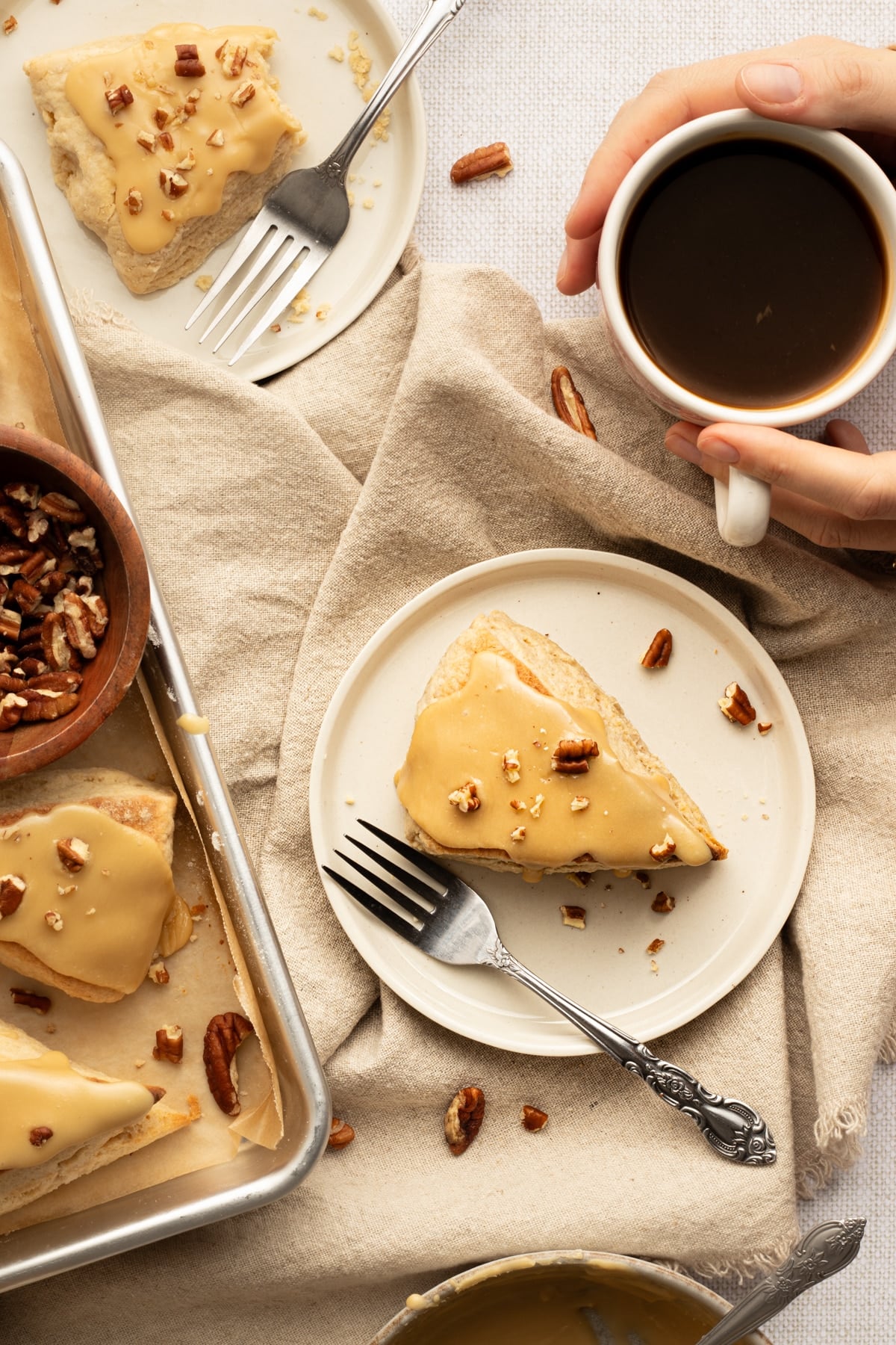 A cozy breakfast scene with glazed maple scones, a mug of black coffee, and scattered pecans on a beige linen backdrop.