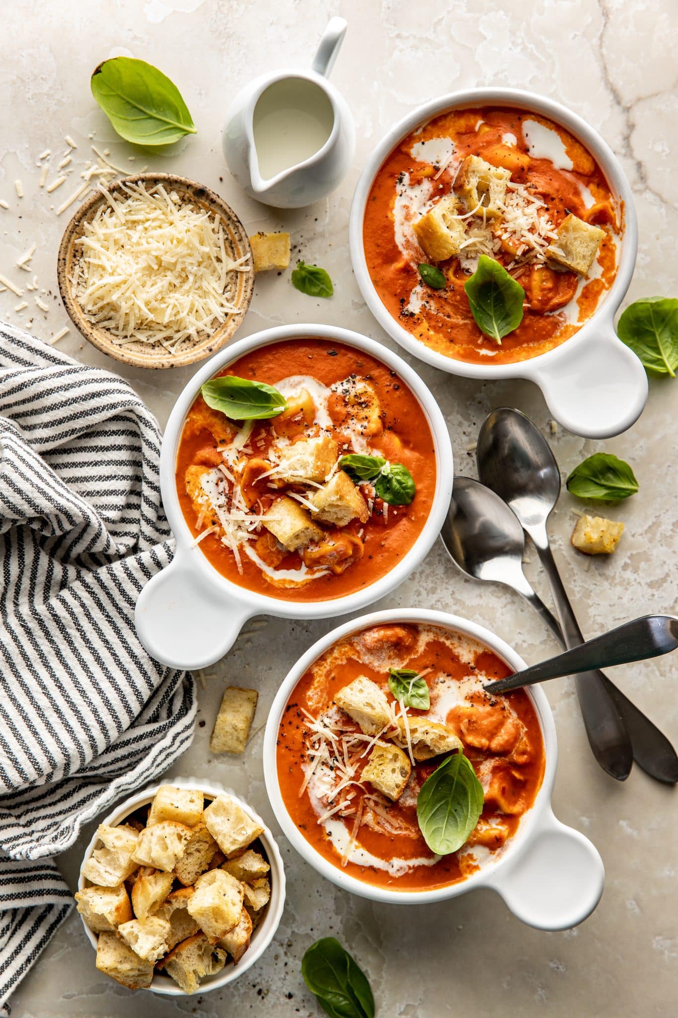 Overhead view of three white bowls filled with creamy tomato tortellini soup, topped with croutons, parmesan, fresh basil, and a drizzle of cream, surrounded by serving spoons and garnishes.

