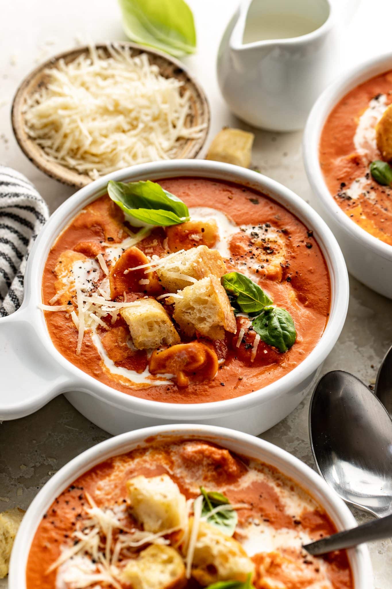 Side angle of a white bowl filled with creamy tomato tortellini soup topped with basil, parmesan, and croutons, with a pitcher of cream and a bowl of shredded cheese in the background.

