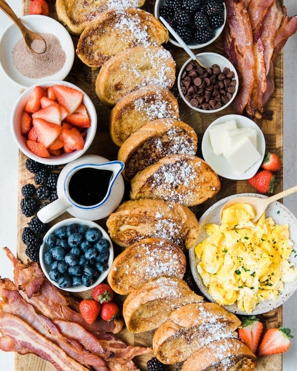 a cutting board filled with french toast, fresh berries, bacon and syrup