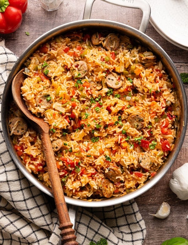 Overhead view of sausage, peppers, and rice skillet garnished with fresh parsley, shown in a stainless steel pan with a wooden spoon.