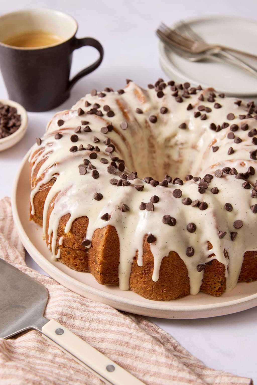 Chocolate chip Bundt cake topped with vanilla bean glaze and mini chocolate chips, displayed on a serving platter with a coffee mug and cake server nearby.