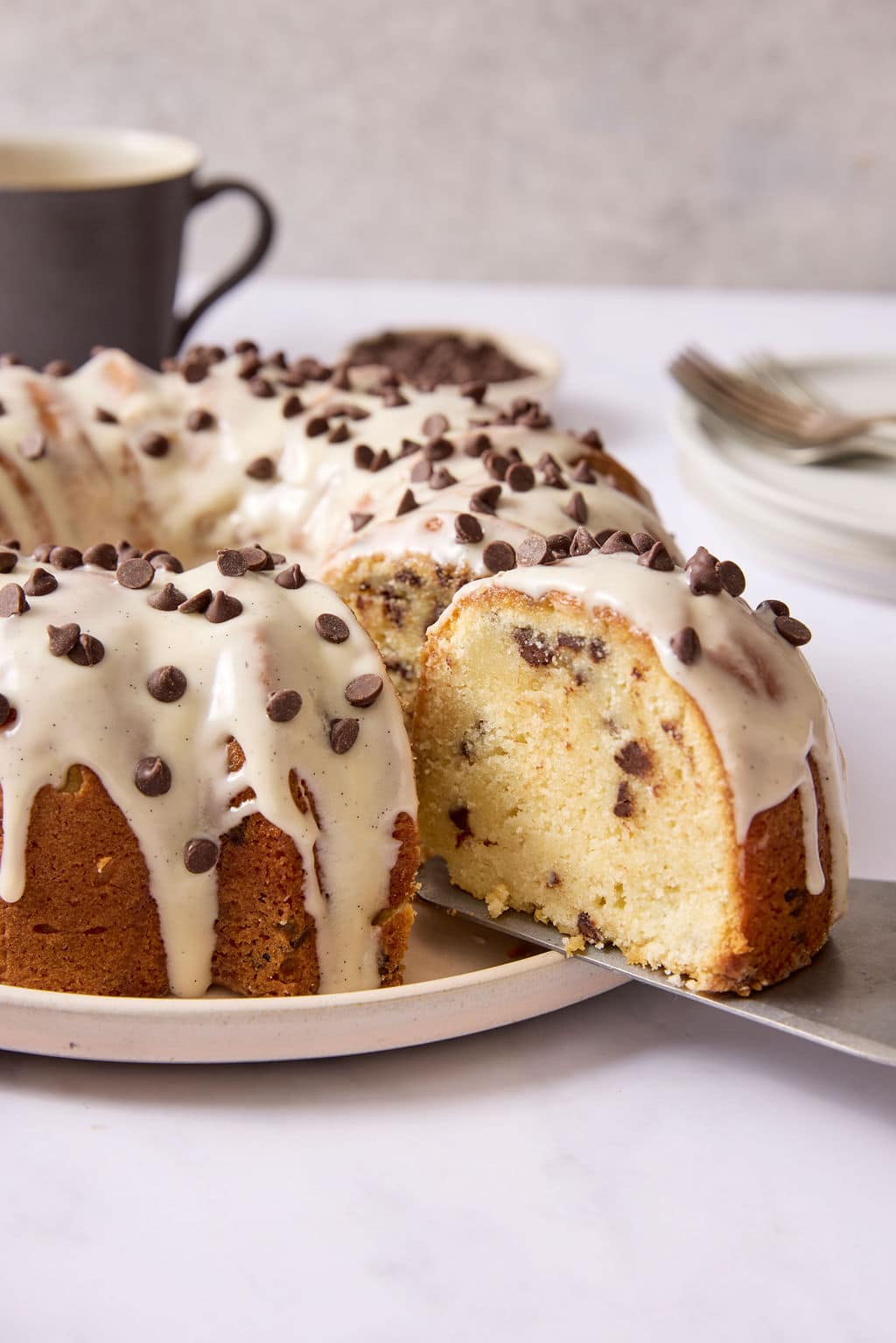 A slice of chocolate chip Bundt cake being lifted from the cake with a server, revealing the moist interior studded with mini chocolate chips and topped with vanilla bean glaze.