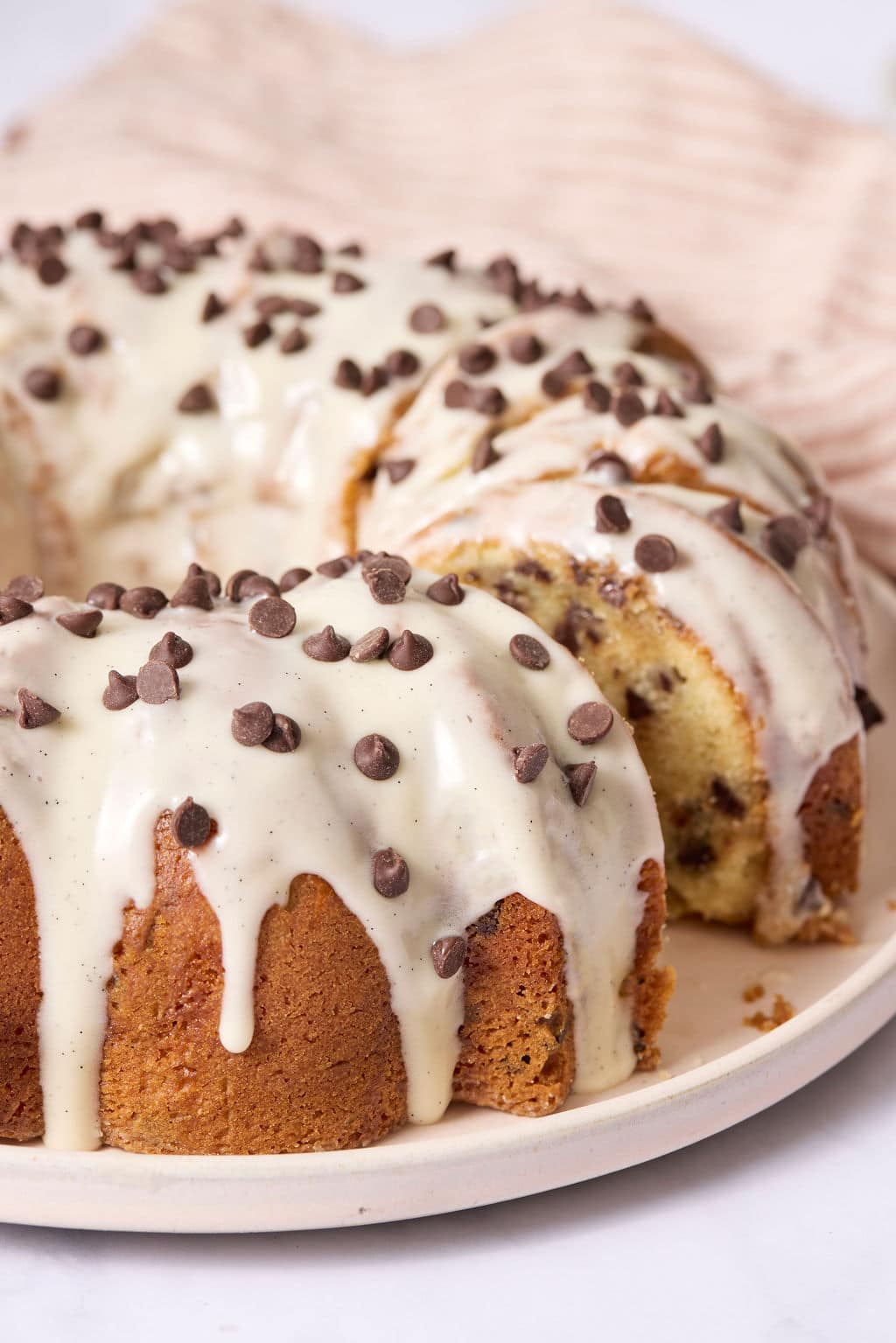 Close-up of a chocolate chip Bundt cake coated in vanilla bean glaze and topped with mini chocolate chips, with a sliced section revealing the tender crumb inside.