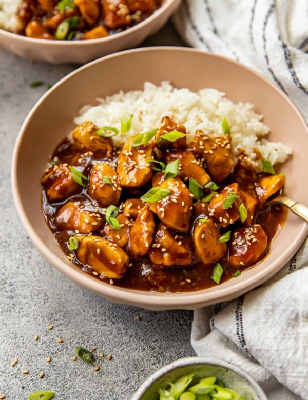 brown bowl with white rice, orange chicken, topped with chopped green onions and sesame seeds