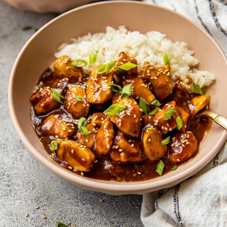 brown bowl with white rice, orange chicken, topped with chopped green onions and sesame seeds