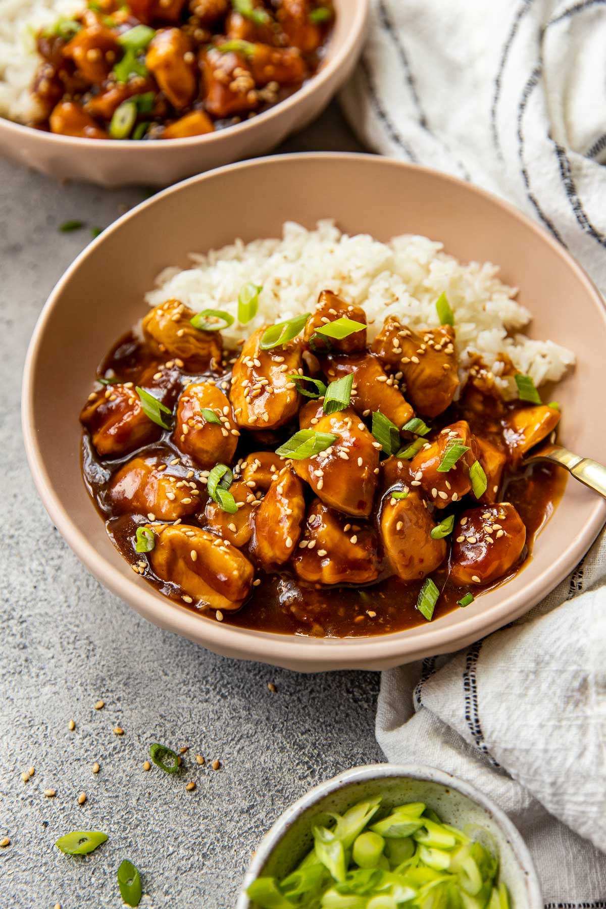 brown bowl with white rice, orange chicken, topped with chopped green onions and sesame seeds