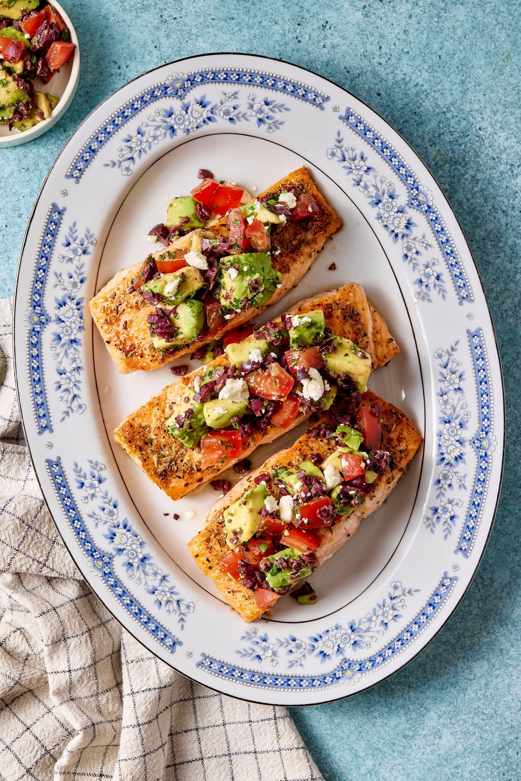 Mediterranean salmon fillets topped with avocado, tomato, olives, and feta arranged on a blue-and-white serving platter.