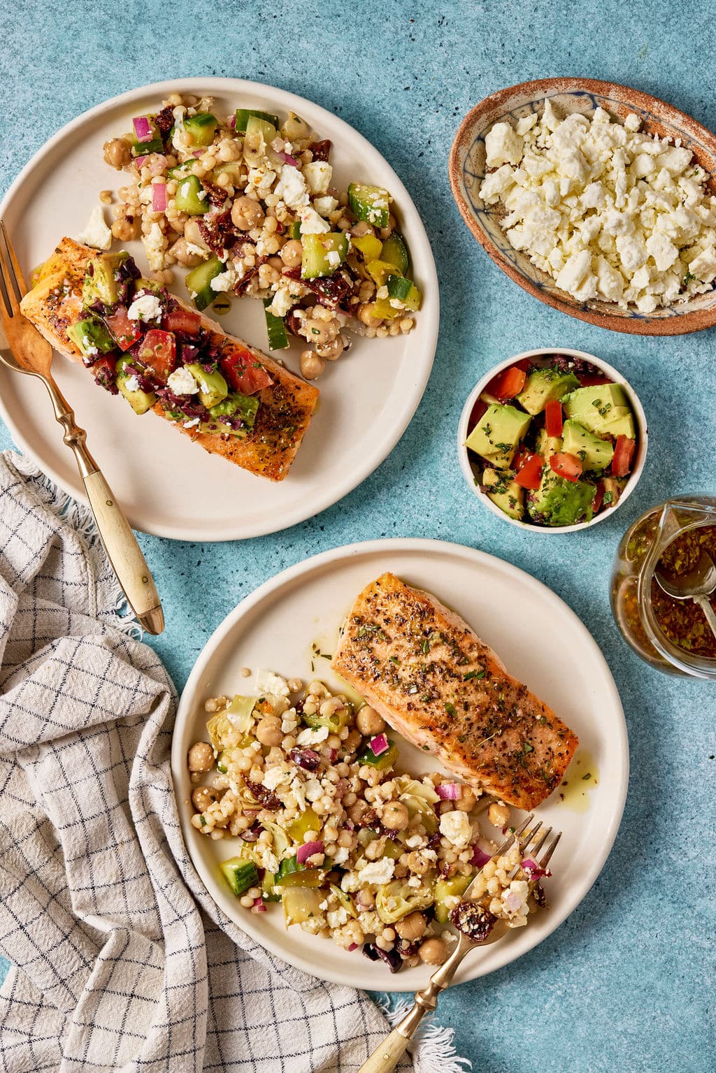 Mediterranean salmon with avocado tomato olive salsa served alongside couscous salad, feta cheese, and vinaigrette on a blue tabletop.