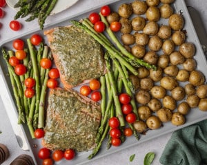 Sheet pan with pesto-coated salmon fillets, asparagus, cherry tomatoes, and seasoned baby Yukon gold potatoes arranged before roasting.
