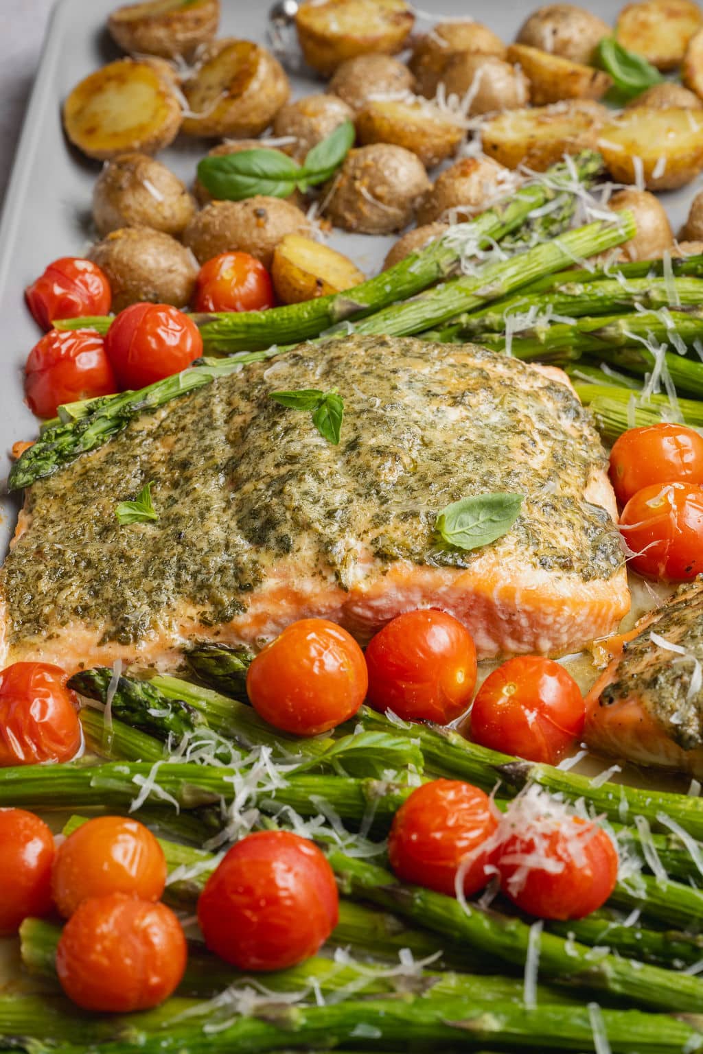 Close-up of sheet pan pesto salmon topped with basil pesto, served with roasted asparagus, blistered cherry tomatoes, crispy baby potatoes, Parmesan cheese, and fresh basil.