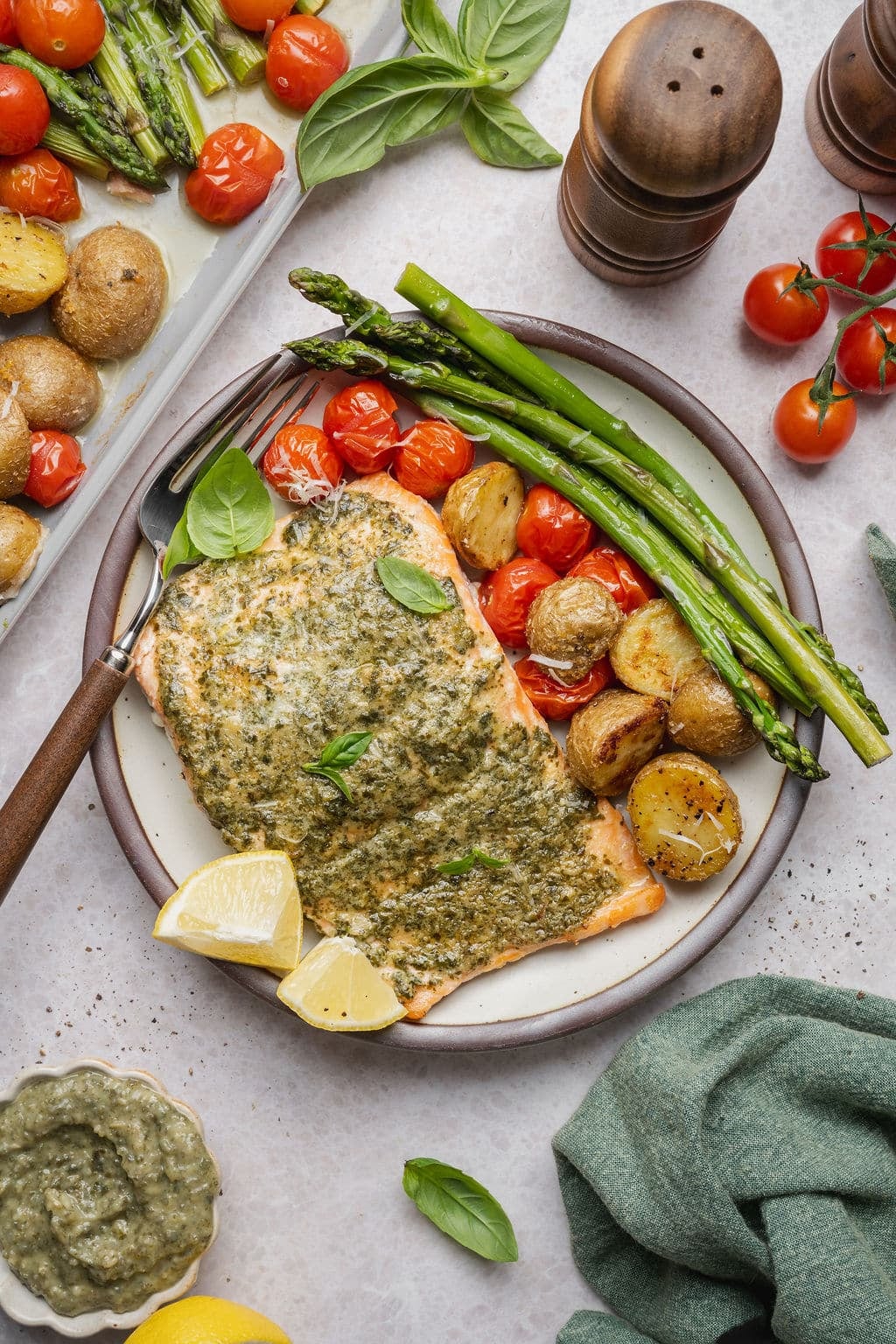 Sheet pan pesto salmon plated with roasted asparagus, crispy baby potatoes, blistered cherry tomatoes, fresh basil, grated Parmesan cheese, and lemon wedges.