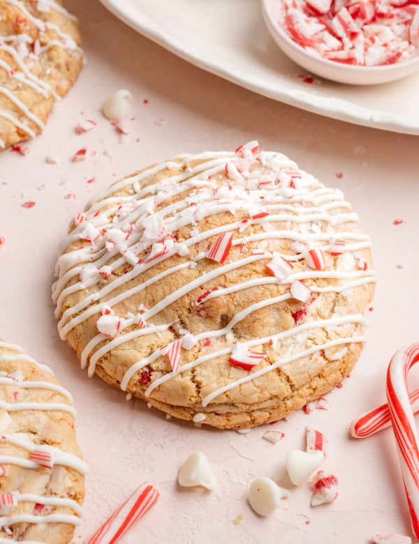 A round white chocolate peppermint cookie covered in white chocolate drizzle and crushed candy cane pieces, surrounded by scattered peppermint crumbs and candy canes.