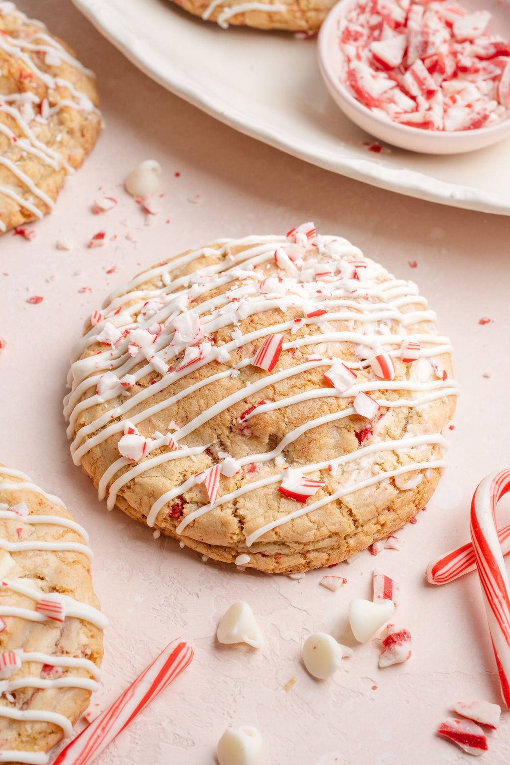 A round white chocolate peppermint cookie covered in white chocolate drizzle and crushed candy cane pieces, surrounded by scattered peppermint crumbs and candy canes.