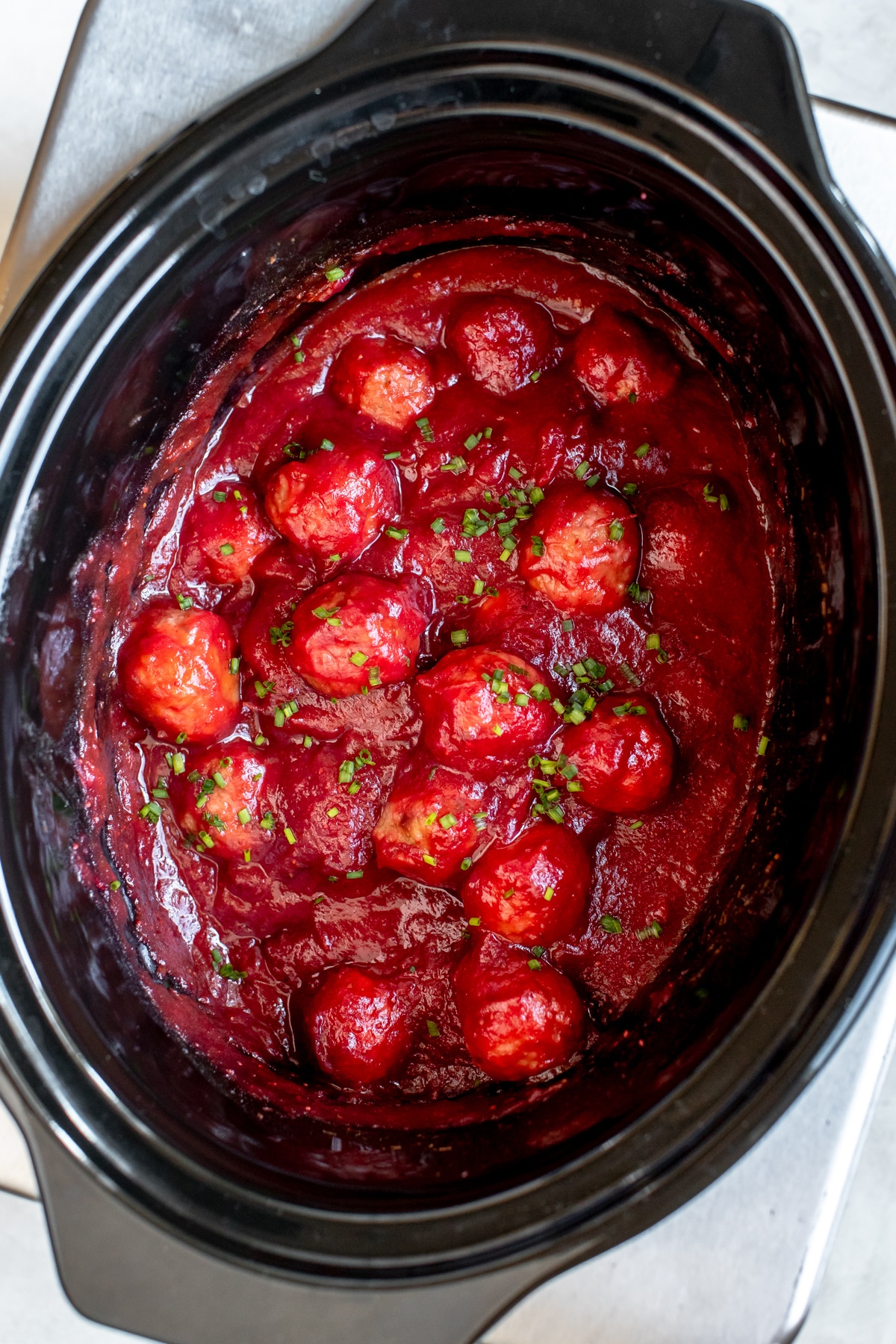 meatballs in a black slow cooker covered in sauce and garnished with chives 