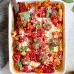 baked meatballs with rigatoni pasta in a white casserole dish topped with basil and parmesan cheese