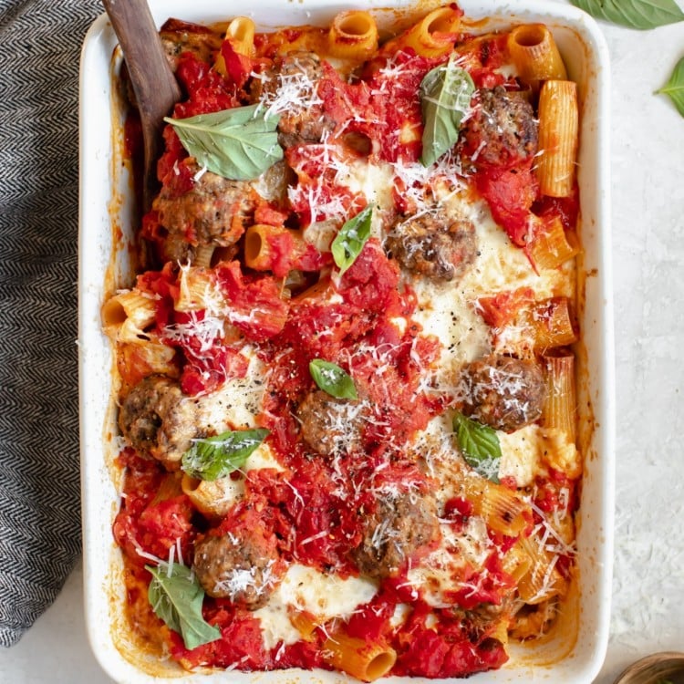 baked meatballs with rigatoni pasta in a white casserole dish topped with basil and parmesan cheese