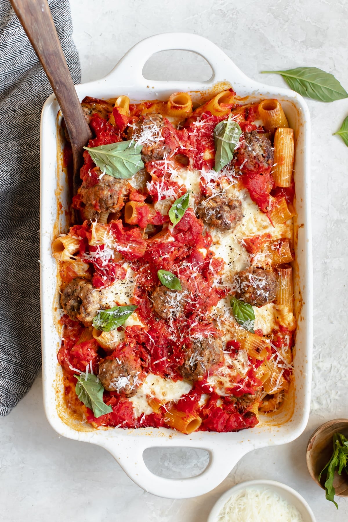 baked meatballs with rigatoni pasta in a white casserole dish topped with basil and parmesan cheese