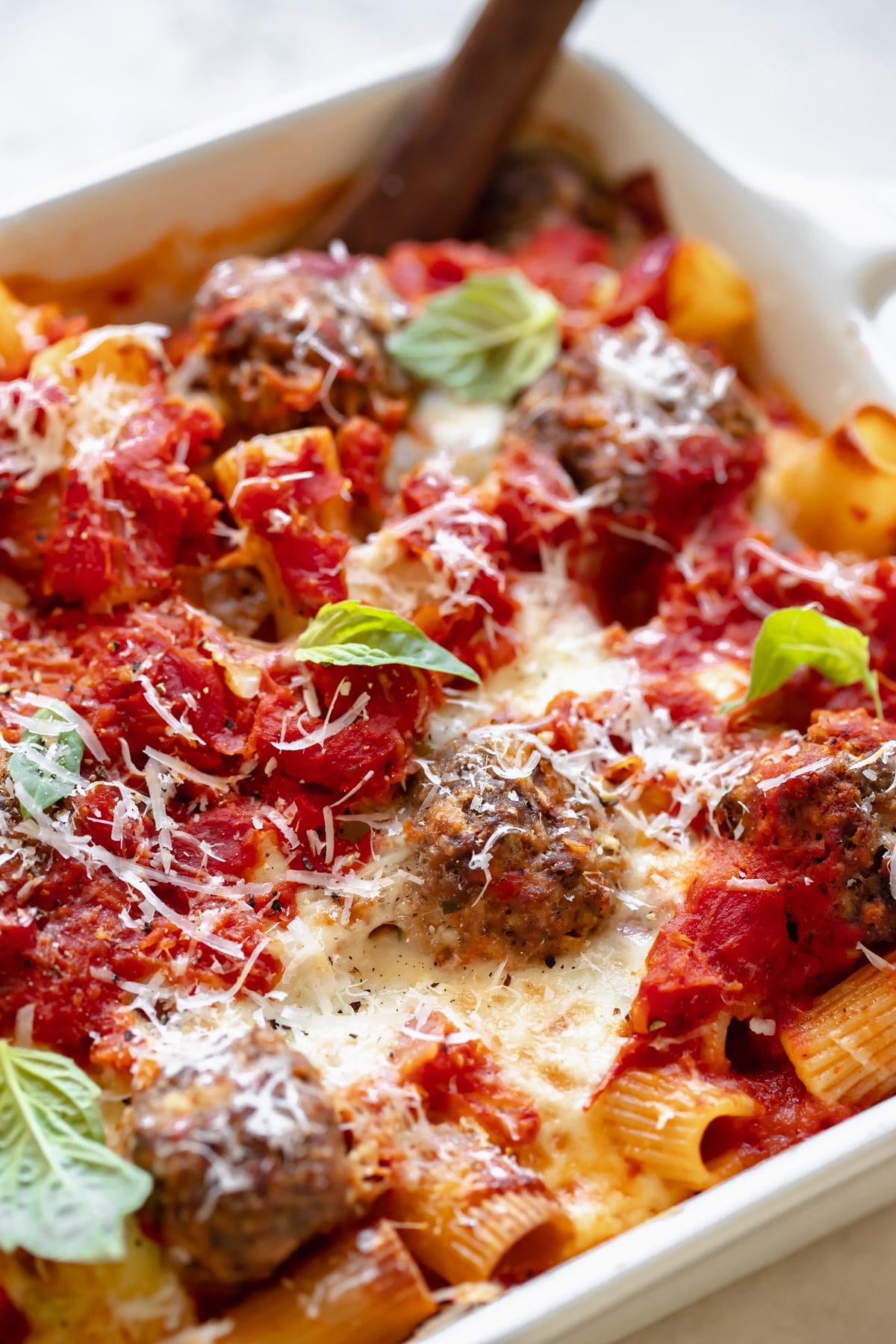 baked meatballs with rigatoni pasta in a white casserole dish topped with basil and parmesan cheese