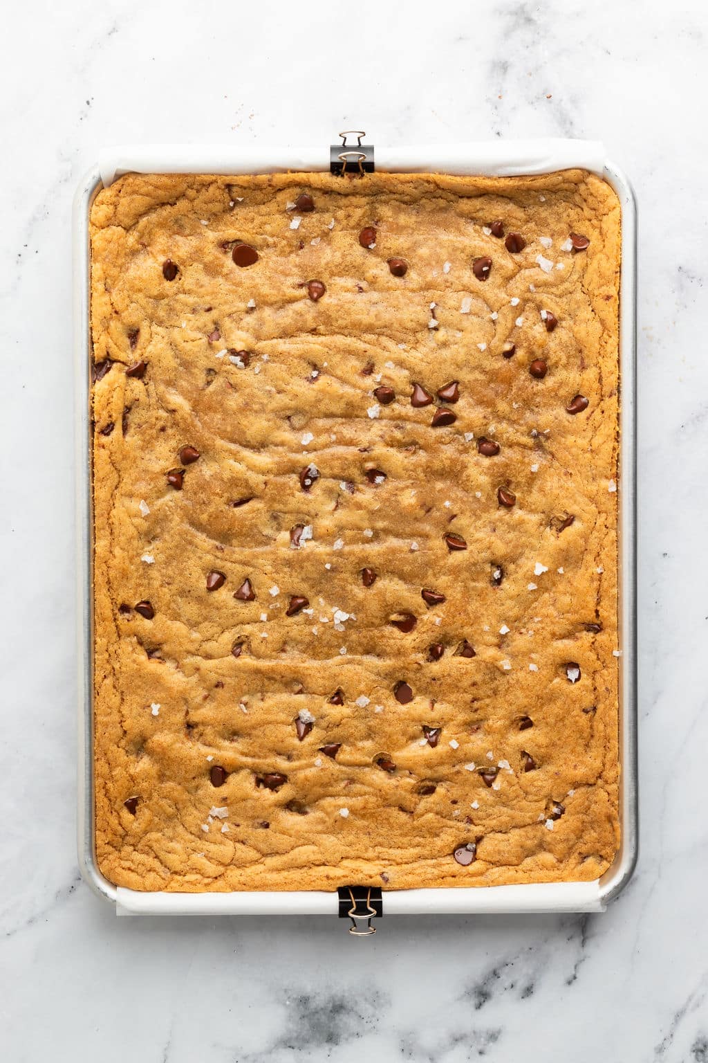 Overhead view of a large rectangular pan of golden brown blondies studded with melty chocolate chips and sprinkled with flaky sea salt. The edges are slightly crisp, and the surface is lightly wrinkled, indicating a soft, chewy center.