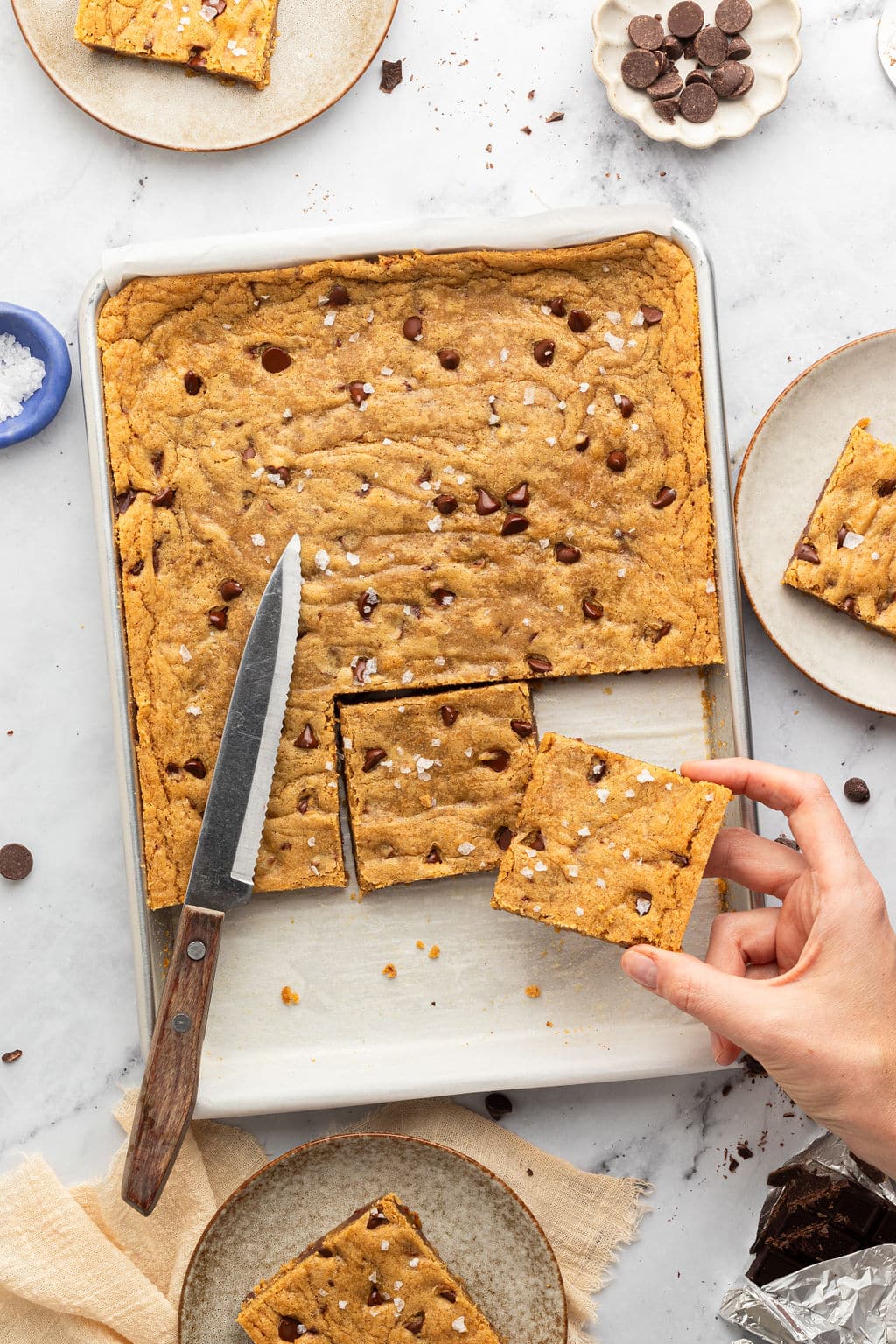 Scene of sliced blondies in a pan, with a hand lifting one square piece. A serrated knife rests on the pan, and scattered plates and chocolate pieces add a cozy, homemade vibe.