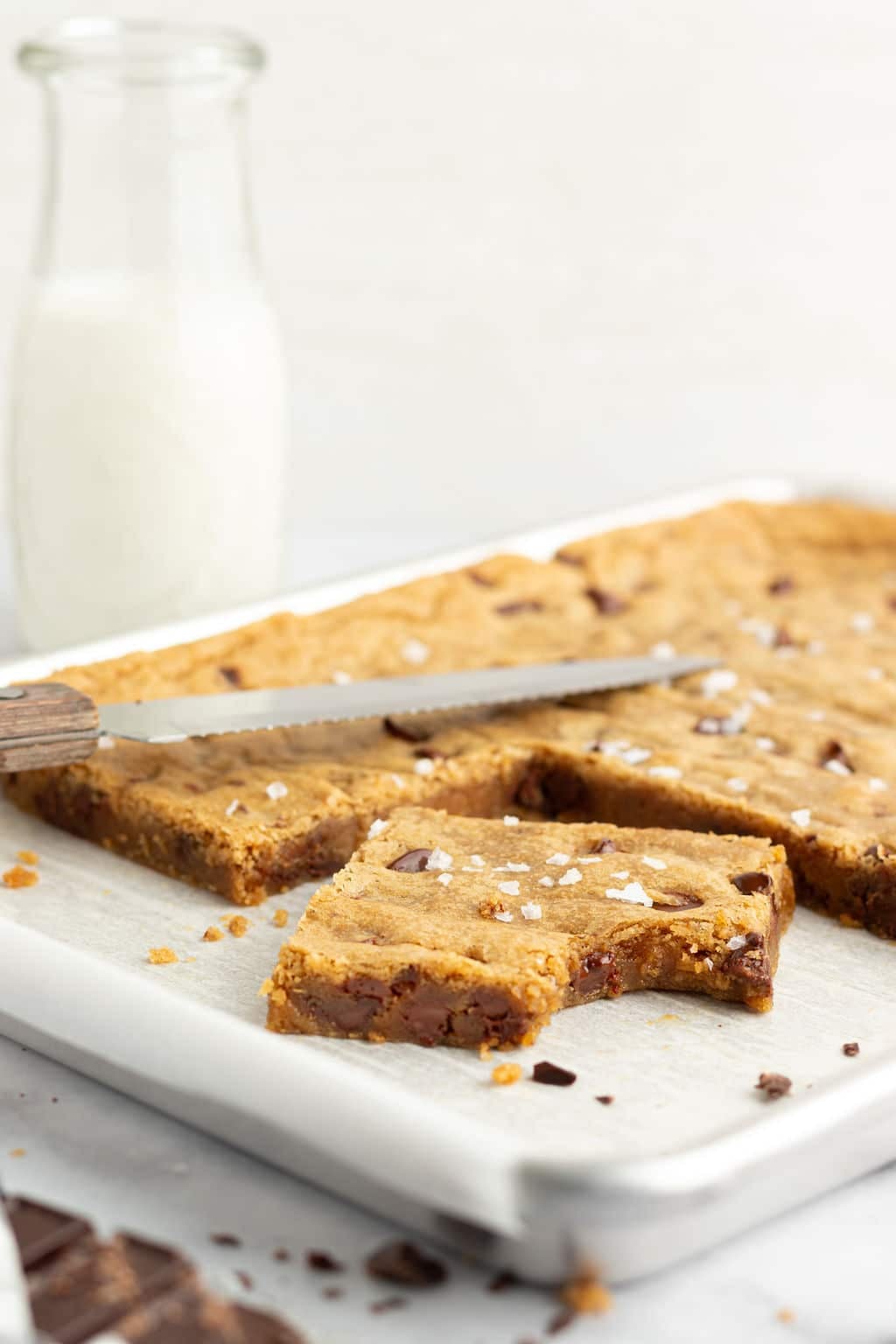 Side-angle of a pan of sliced blondies, with a serrated knife and one blondie missing a bite in the foreground. Flaky salt and gooey chocolate chips are visible in the chewy center.