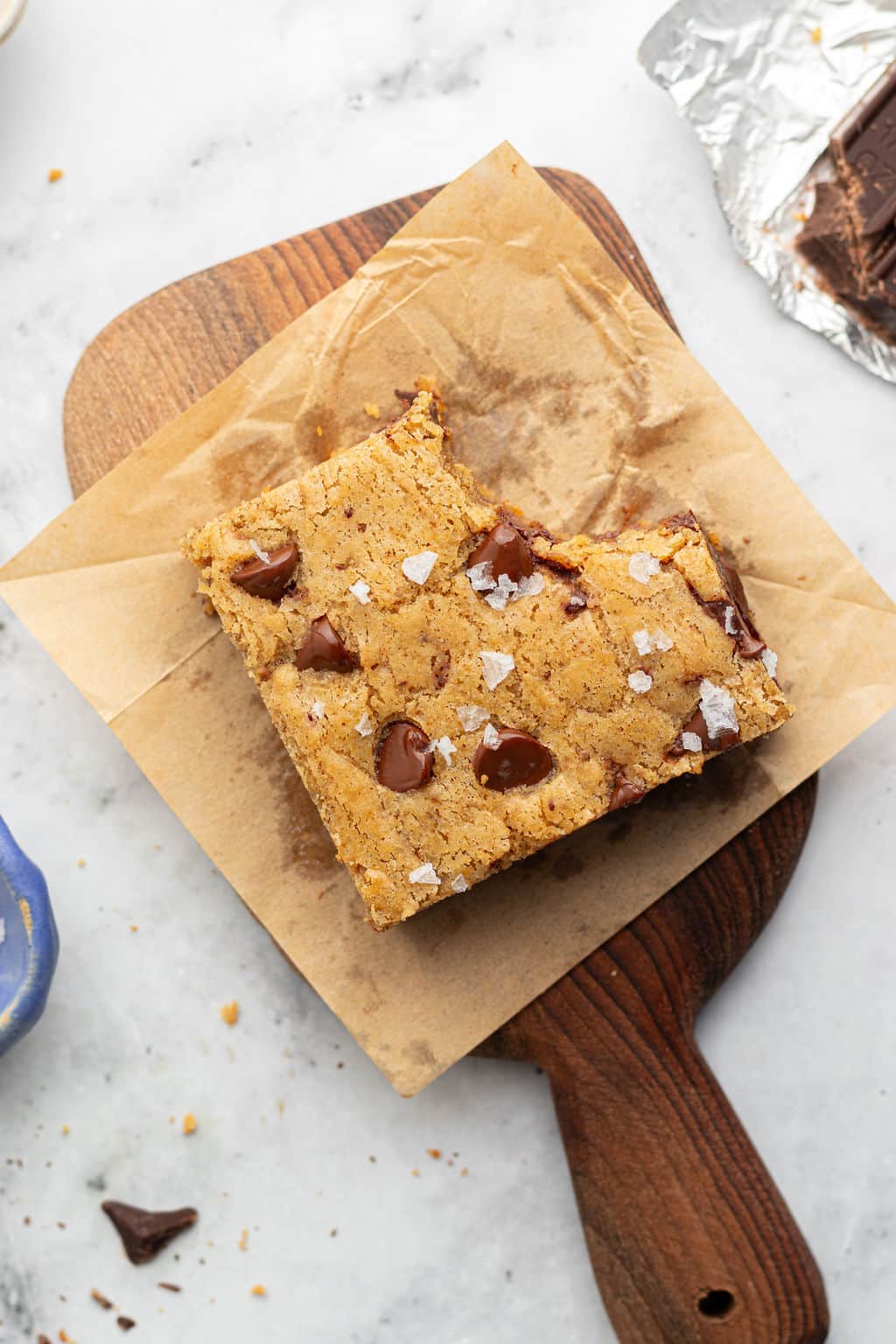 Overhead view of a single square blondie on parchment paper atop a wooden board. A bite has been taken from the top corner, exposing a soft and chewy interior with chocolate chips and sea salt.