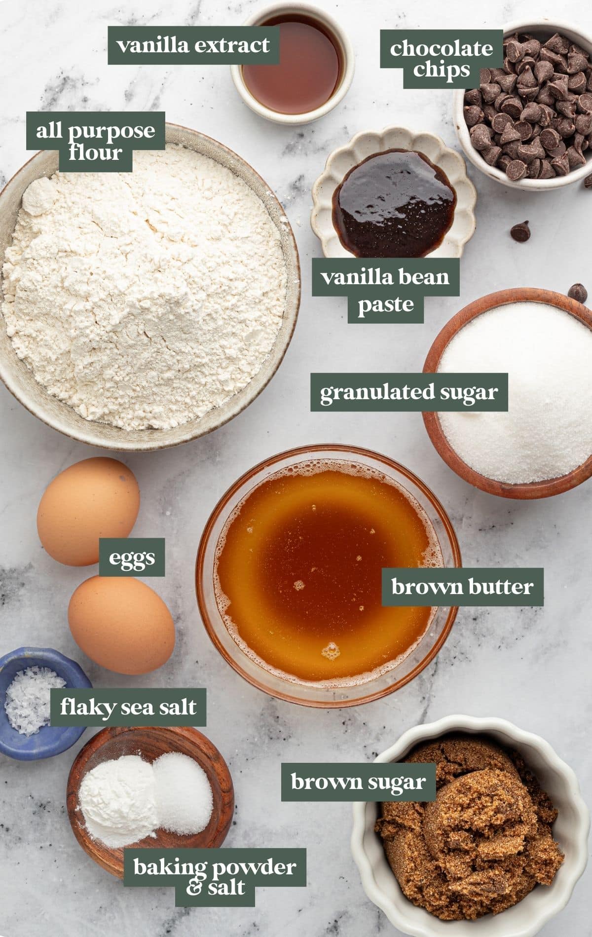 Overhead image of labeled ingredients for brown butter blondies arranged on a light marble surface. Items include a bowl of all-purpose flour, a small dish of vanilla extract, a bowl of chocolate chips, a small scalloped dish of vanilla bean paste, a bowl of granulated sugar, a bowl of rich golden brown butter, a bowl of brown sugar, two whole eggs, a pinch dish of flaky sea salt, a small wooden dish containing baking powder and salt, all neatly styled for baking preparation.