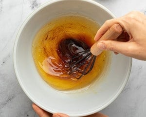 A hand uses a whisk to stir warm brown butter and vanilla bean paste in a white mixing bowl on a marble surface.
