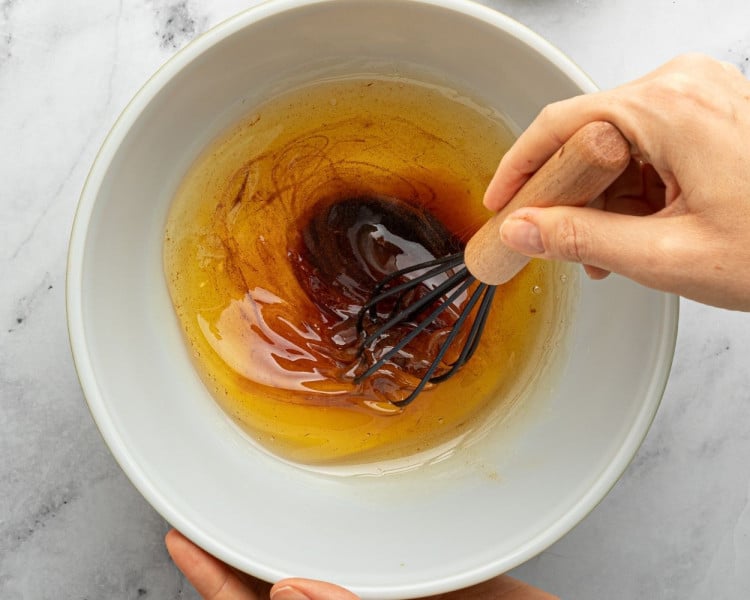 A hand uses a whisk to stir warm brown butter and vanilla bean paste in a white mixing bowl on a marble surface.