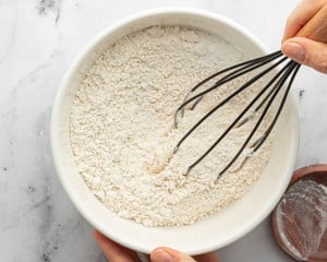 Dry ingredients, including flour, baking powder, and salt, being whisked together in a white bowl by hand.