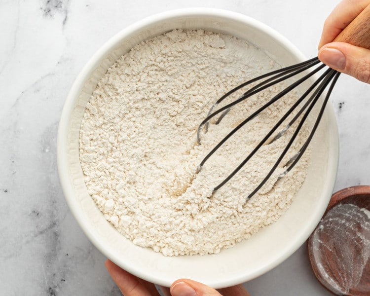 Dry ingredients, including flour, baking powder, and salt, being whisked together in a white bowl by hand.