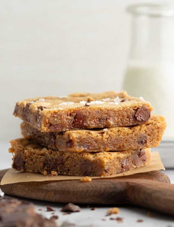 Side view of three stacked blondie bars on a wooden board with parchment paper. The blondies are thick, golden, and studded with large chocolate chunks, with a bottle of milk in the soft-focus background.