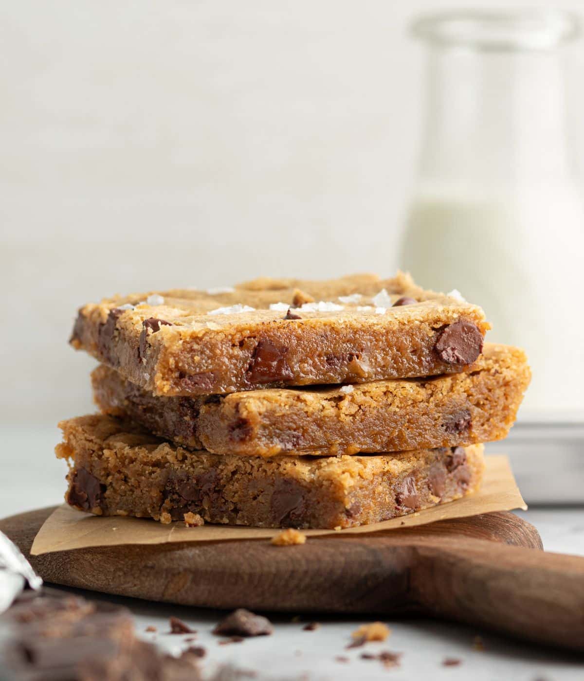 Side view of three stacked blondie bars on a wooden board with parchment paper. The blondies are thick, golden, and studded with large chocolate chunks, with a bottle of milk in the soft-focus background.