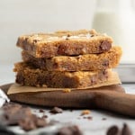 Side view of three stacked blondie bars on a wooden board with parchment paper. The blondies are thick, golden, and studded with large chocolate chunks, with a bottle of milk in the soft-focus background.