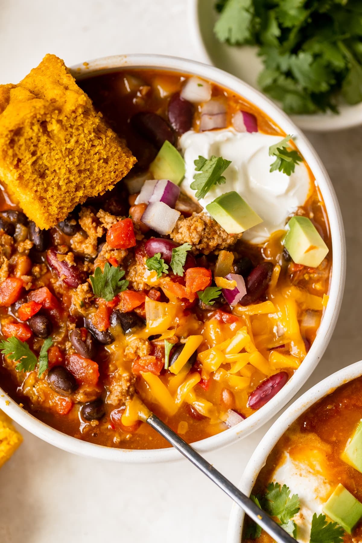 a white bowl filled with chili made with beans and garnished with red onion, cheese, cilantro, sour cream and avocado. 